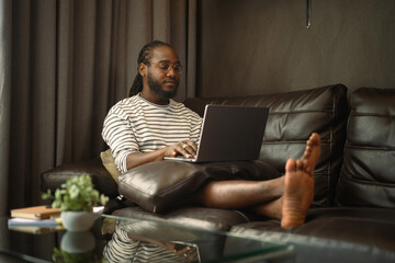Casual young African man working with laptop on couch at home