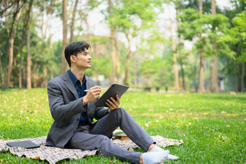 Portrait of handsome businessman sitting grass holding digital tablet and looking away