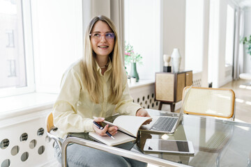 A cheerful woman in glasses makes notes in a notebook beside her laptop in a luminous, stylishly...