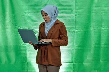 shocked surprised woman wearing hijab and blazer standing and holding laptop computer isolated over...