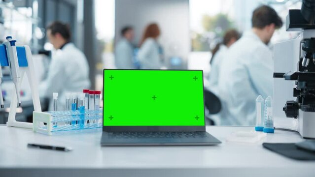 Laptop Computer with Green Screen Chromakey Template Standing on a Desk in a Science Laboratory Next to a Microscope and Test Tubes with Genomics Samples. Scientists Working in the Background