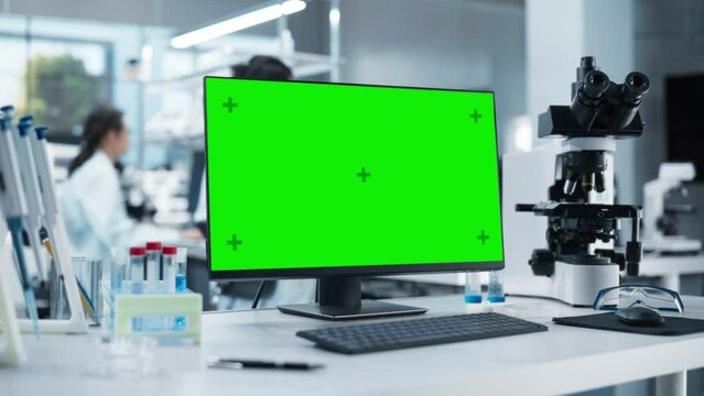 Desktop Computer with Green Screen Mock Up Display Standing on a Table in a Science Laboratory Next to a Microscope and Test Tubes with Biological Samples. Anonymous People Working in the Background