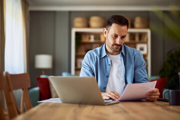 A focused adult freelance man reading a document and sitting in front of a laptop in a home office.