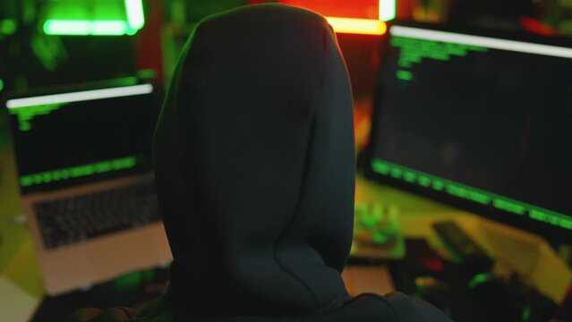 Hacker writing code cracking computer. Man programmer in hoodie cracks system, using computer in dark room back view. Hacker attack, technology, virus infected software, cyber information concept.