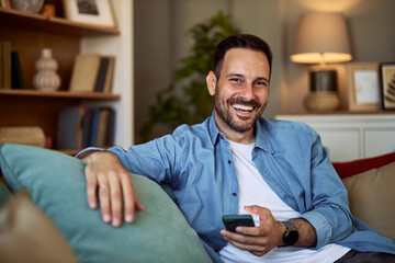 Portrait of a smiling young adult man casually sitting on a couch and holding a cellphone. - obrazy, fototapety, plakaty