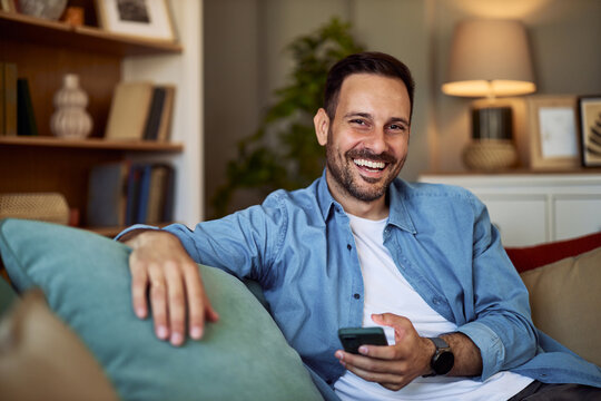Portrait of a smiling young adult man casually sitting on a couch and holding a cellphone.