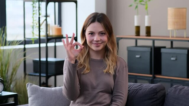 Positive and friendly Caucasian young woman showing ok sign in agreement and smiling at camera at home. Approve and accept gesture, symbol.