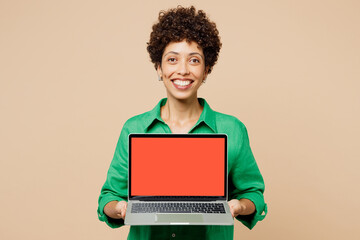Young IT woman of African American ethnicity wears green shirt casual clothes hold use work on blank screen area laptop pc computer look camera isolated on plain beige background. Lifestyle concept.