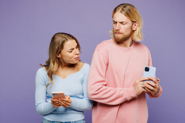 Young sad indignant suspicious couple two friends family man woman wear pink blue casual clothes together hold in hand use try to check boyfriend mobile cell phone isolated on plain purple background.