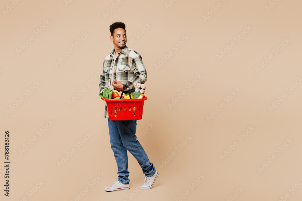 Poster Full body side view young smiling happy cheerful man wearing grey shirt hold red basket bag with food products look aside isolated on plain beige background. Delivery service from shop or restaurant.