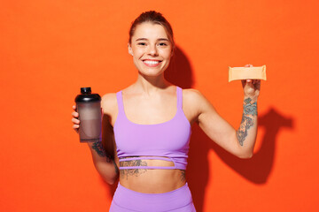 Young happy fitness trainer instructor sporty woman sportsman wear purple top clothes in home gym eat protein bar hold cocktail shake isolated on plain orange background Workout sport fit abs concept