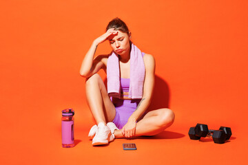 Full body sad young fitness trainer woman sportsman wears top shorts purple clothes in home gym sit...