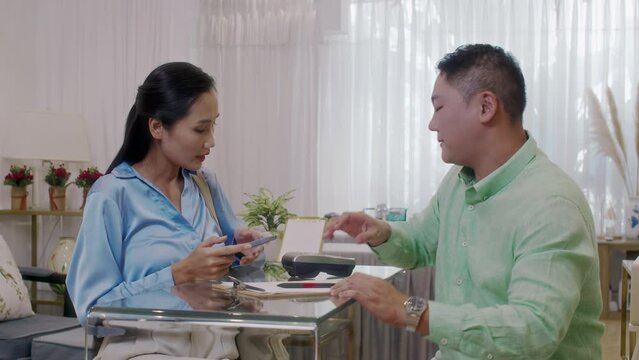 Medium shot of young Asian woman paying with smartphone for procedure to male spa salon receptionist