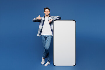 Full body smiling young man he wear shirt white t-shirt casual clothes big huge blank screen mobile cell phone smartphone with area show thumb up isolated on plain blue background. Lifestyle concept.