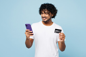 Young Indian man wear white t-shirt casual clothes using mobile cell phone hold credit bank card do online shopping order delivery book tour isolated on plain blue cyan background. Lifestyle concept. - obrazy, fototapety, plakaty
