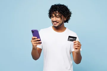 Tuinposter Young Indian man wear white t-shirt casual clothes using mobile cell phone hold credit bank card do online shopping order delivery book tour isolated on plain blue cyan background. Lifestyle concept. © ViDi Studio
