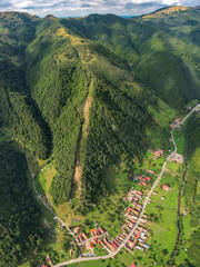 Aerial vertical panorama above a village located inside a mountainous area, along a river. The river winds near beech forests, and meadows. Clouds are casting their shadows on the forests. Carpathia