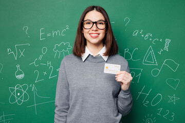 Young happy smart teacher woman wear grey casual shirt glasses hold credit bank card isolated on plain green wall white chalk blackboard background studio. Education in high school college concept.