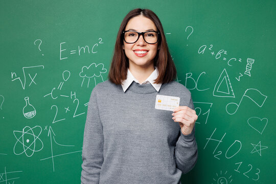 Fototapeta Young happy smart teacher woman wear grey casual shirt glasses hold credit bank card isolated on plain green wall white chalk blackboard background studio. Education in high school college concept.