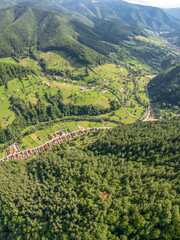 Aerial vertical panorama above a village located inside a mountainous area, along a river. The river winds near beech forests, and meadows. Countryside landscape, Carpathia, Romania.