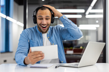Happy and surprised young muslim man sitting in the office at the table wearing headphones and looking enthusiastically at the tablet screen while holding his head with his hand