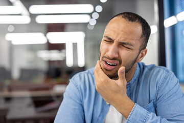 Young hispanic man suffering from a toothache sits in a modern office and grimacing, holding his hand to his cheek. Close-up photo - obrazy, fototapety, plakaty