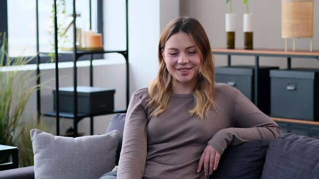 Portrait of a happy and smiling Caucasian young woman sitting on sofa at home and looking at camera. Relaxation on leisure time. Women femininity concept.