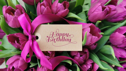 Happy birthday text on label in front of purple flowers - tulips.	