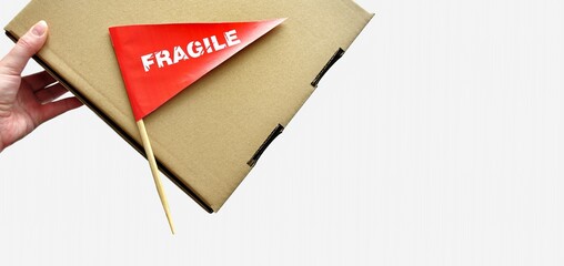 One cardboard packaging box in hands on monochrome background. Tiny red paper flag with the warning 'Fragile' as a label, sticker. The concept of packing and shipping fragile items