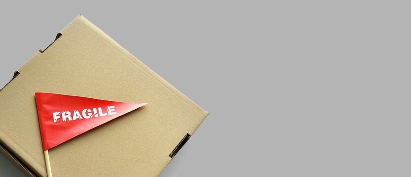 One cardboard packaging box on monochrome background. Tiny red paper flag with the warning 'Fragile' as a label, sticker. The concept of packing and shipping fragile items