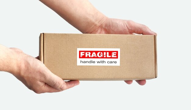 One rectangular cardboard box in hands for fragile items packing on a plain light background. An adhesive red warning label, sticker for packaging 'Fragile, Handle with care, Thank you'