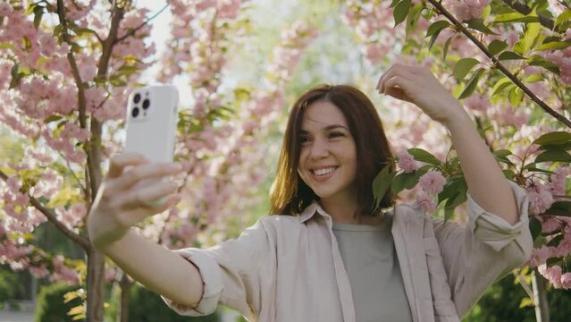 A happy young woman makes a selfie on the background of a tree that blooms with pink flowers. Selfie on a walk, blog, happy moments on camera. Spring flowers of cherry or sakura blossoms on background