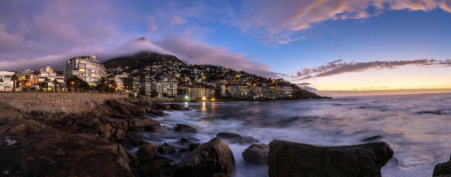 Panoramic view of Bantry Bay city lights at sunset on Sea Point Promenade with dramatic low cloud rolling over Lion&rsquo;s Head mountain in background, Saunder's Rock beach, Cape Town, South Africa.