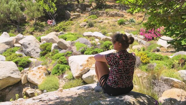 Woman, young female sitting on rock under tree shade on Ikaria island, Greece longevity blue zone alone with nature landscape view meditating