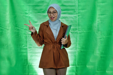 beautiful young Asian woman wearing hijab, glasses and blazer holding laptop and making peace sign...