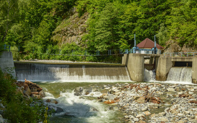 A small power plant with its concrete dam built on a mountain river. The water is overflowing the...