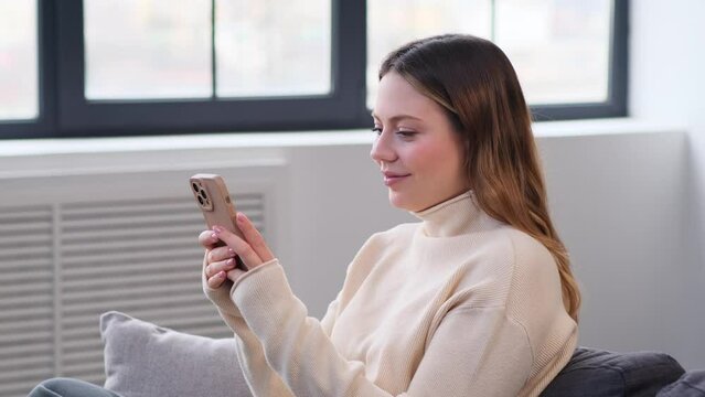 Smiling Caucasian young woman chatting, texting in online social media or browsing internet and laughing using smartphone at home.