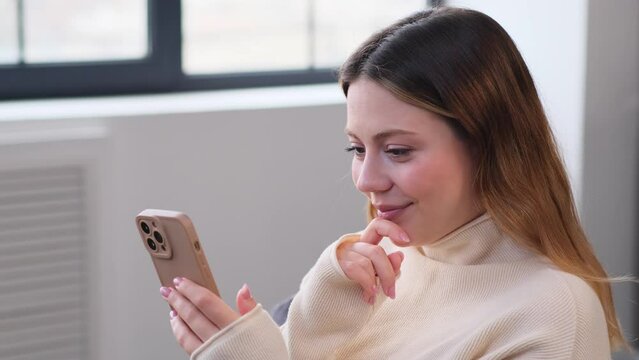 Portrait of a happy Caucasian young woman relaxing online with phone at home. Messaging in social media app, reading news, browsing internet.