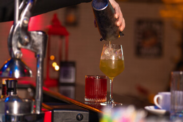 Close-up of the bartender carefully pours a ready-made cocktail from a metal shaker into a glass with ice behind the bar. - obrazy, fototapety, plakaty