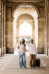 Two young cheerful women pulling a suitcase and walking together through a city during a weekend...