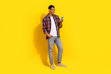 Full size photo of good mood man dressed plaid shirt jeans trousers look at smartphone read email isolated on yellow color background