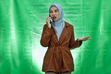 angry young Asian woman wearing hijab and blazer talking on mobile with raised hands over green...