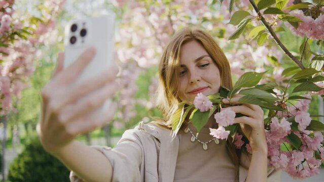 A happy young red-haired woman makes a selfie on the background of a tree that blooms with pink flowers. Selfie on a walk, blog, happy moments on camera. Spring flowers of cherry or sakura blossoms on
