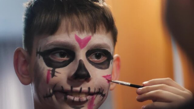 a 9-year-old boy paints his face with carnival makeup in front of a mirror for Halloween or the Day of the Dead. a child prepares for a holiday in a mystical costume