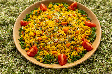 Millet groats cooked with turmeric and vegetables, decorated with chopped chives and tomato quarters, then sprinkled with alfalfa sprouts on a plate. Alfalfa sprouts make the background.