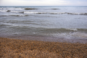 A beach with small shells and the sea. The coast of the Caspian Sea. Cool weather on the coast. A walk along the seashore. The healing sea air. Waves on the surface of the water. Cloudy weather at sea