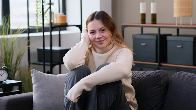 Portrait of a cool and friendly Caucasian young woman sitting on sofa, looking and smiling at camera, at home living room. Satisfied from resting and weekend.