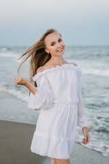 Fototapeta na wymiar Female stand on the beach ocean and enjoy summer day on vacation. Portrait of a beautiful girl in a white dress. Happy woman go on seashore. Girl walks in water with big waves on sand sea in evening.