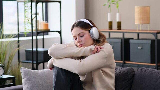 Melancholy and hopeless Caucasian young woman listening music in headphones, sitting on sofa at home. Sadness and apathy during weekend.