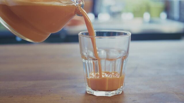 Woman pouring fresh, freshly pressed carrot juice from a glass jar into a glass standing on a wooden table. Defocused brightly lit kitchen in the background. High quality 4k footage